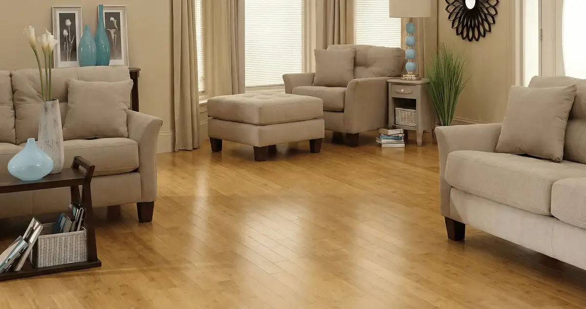 A living room with professionally installed bamboo flooring, beige sofas, a matching ottoman, a side table with a blue lamp, vases, framed pictures, and large windows with blinds and curtains in Reno, NV